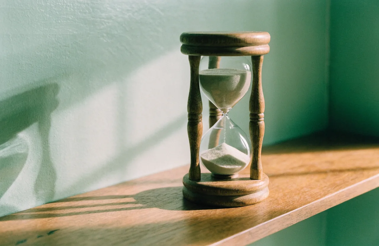 A wooden hourglass with pale sand on a wooden shelf against a muted sage wall.